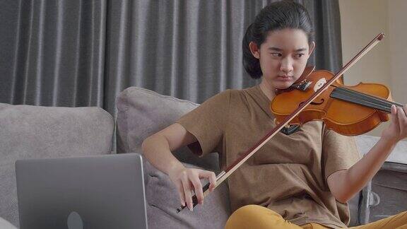 迷人的亚洲少女在冠状病毒封锁的情况下待在家里用笔记本电脑与老师在线练习小提琴并学习小提琴弹奏古典乐器的年轻女子
