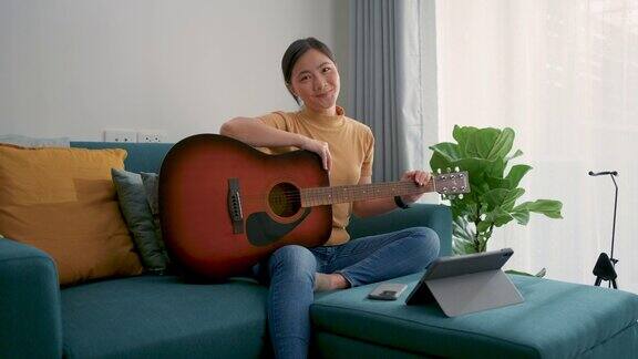 一个亚洲女人拿着吉他坐在沙发上在家里的客厅里用平板电脑学习吉他微笑着看着镜头