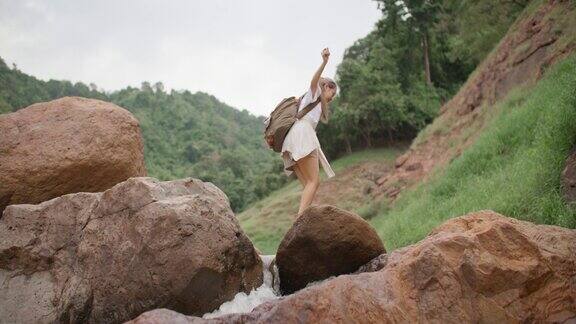 年轻的女旅行者在森林小径山上的岩石上跳过小溪