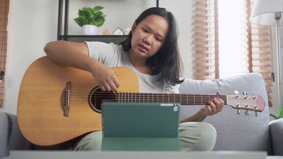 一名年轻的亚洲女子坐在沙发上用家里的笔记本电脑在网上学习吉他