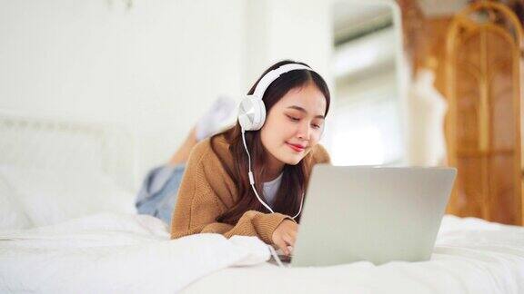 快乐美丽的年轻亚洲女人工作的电脑笔记本电脑躺在床上的房子她在做视频会议、网络会议或者戴着耳机听音乐
