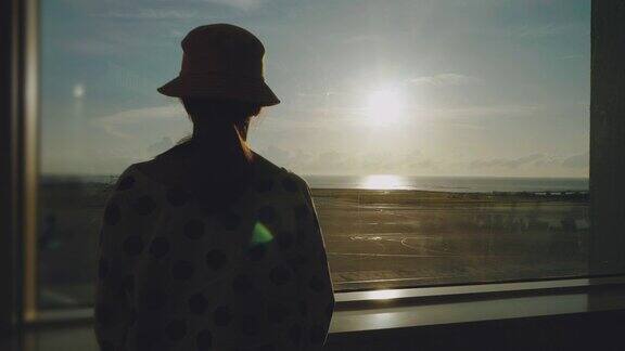 年轻的亚洲女子站在机场的窗口观察日落的飞机