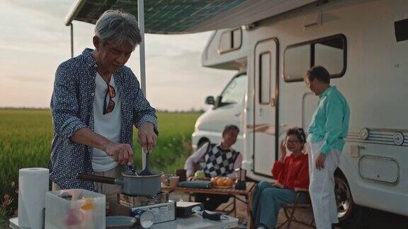 亚洲华裔老人在周末休闲活动期间在稻田边的露营车旁为他的高级朋友做饭准备晚餐