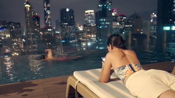晚上一名亚洲男子在一座高层建筑的游泳池里游泳一名女子拿着手机坐在那里俯瞰整个城市