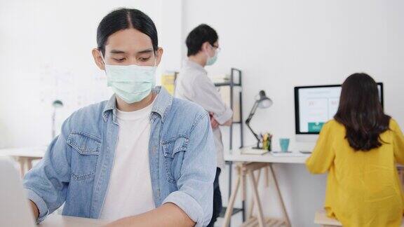 在新常态下亚洲商人企业家戴着医用口罩进行社交距离以预防病毒同时在办公室使用笔记本电脑工作