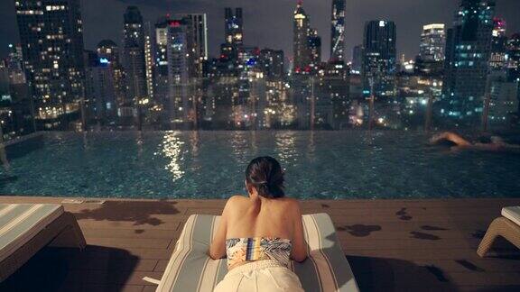 晚上一名亚洲男子在一座高层建筑的游泳池里游泳一名女子拿着手机坐在那里俯瞰整个城市