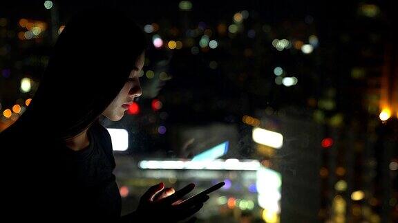 年轻的亚洲女性在夜晚使用手机以城市的灯光为背景