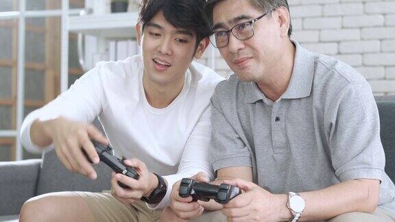 年长的亚洲男人在退休年龄有乐趣玩视频游戏机与他的儿子在他的舒适的家