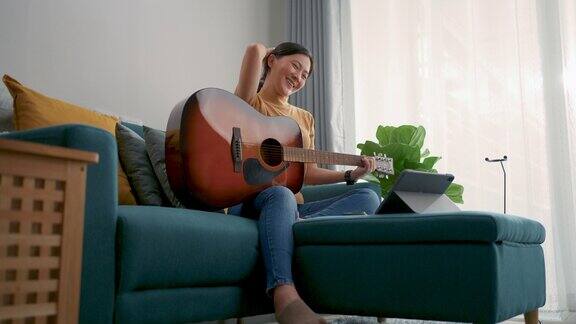 一个亚洲女人拿着吉他坐在沙发上在家里的客厅里用平板电脑学习吉他
