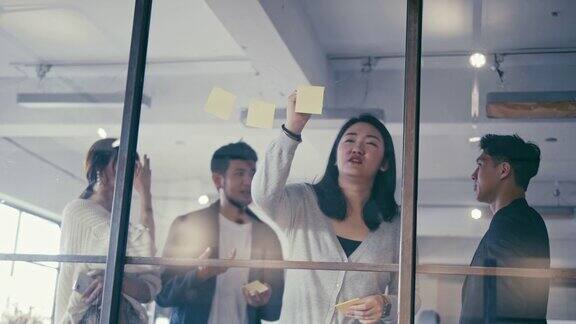一群亚洲的办公室职员在玻璃墙上贴着不干胶的便条头脑风暴(慢镜头)