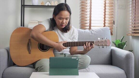 亚洲妇女坐在沙发上练习弹木吉他从网上教程在家里学习以技能提升为理念的周末活动