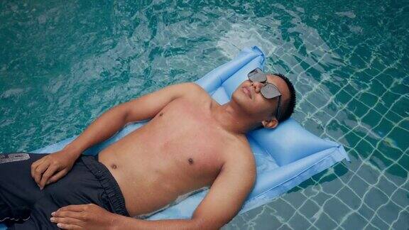 戴着墨镜的年轻男子正在酒店游泳池里漂浮的蓝色充气床上放松