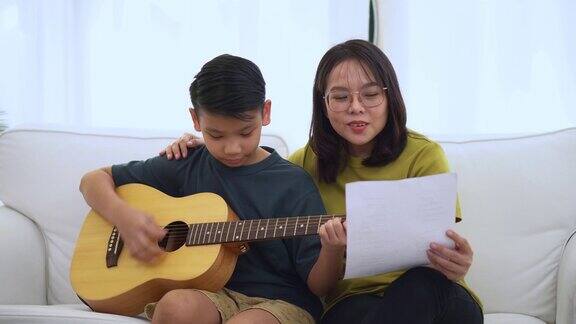 亚洲母亲拥抱儿子亚洲男孩弹吉他母亲拥抱在沙发上感到感激和鼓励幸福的家庭观念学习有趣的生活方式爱的亲情纽带