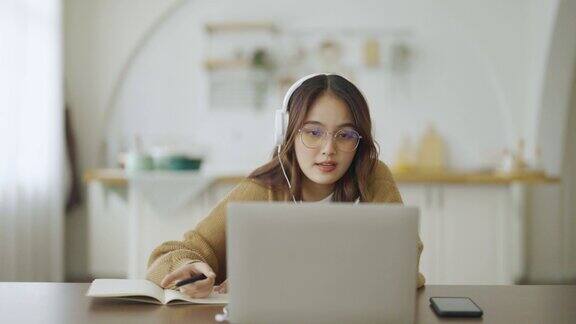 年轻的亚洲女性戴着眼镜和耳机在家里的电脑笔记本电脑上工作在家办公视频会议视频通话学生在线学习