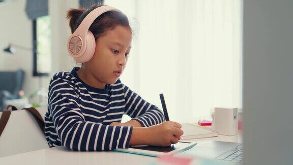 亚洲学步女孩戴着耳机坐在书桌前用笔记本电脑使用魔笔专注写作做作业手指数数数学在线学习课程从笔记本电脑在家里