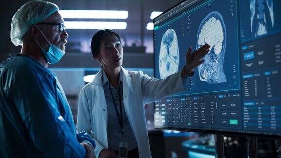 医院研究实验室会议:亚洲女性神经科学家与高加索男性神经外科医生讨论电视屏幕上的CT脑部扫描穿着手术服准备手术的男子