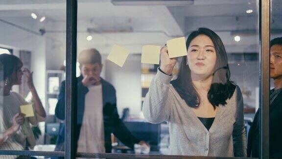 一群亚洲的办公室职员在玻璃墙上贴着不干胶的便条头脑风暴(慢镜头)