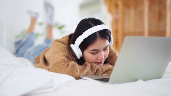 快乐美丽的年轻亚洲女人在电脑上工作躺在床上她开视频会议或者戴着耳机听音乐
