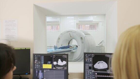 两名合格的放射科医生检查躺在CT或MRI扫描仪下的女性病人计算机屏幕显示诊断结果图像透过现代诊所的玻璃窗观看