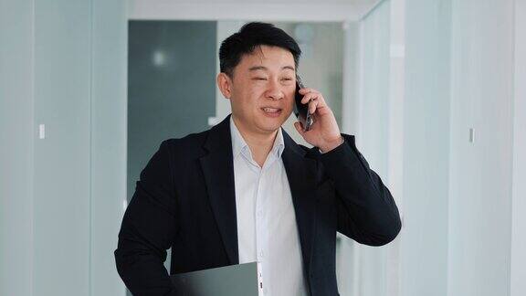 富有魅力的亚洲程序员拿着现代笔记本电脑打电话开朗的商人在办公室走廊打电话快乐的商人肖像在室内打电话