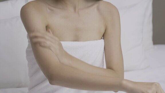 近距离的女人在毛巾上坐在床上使用清爽霜或身体乳液在她的手和肩膀
