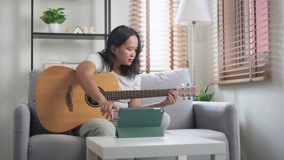 亚洲妇女坐在沙发上练习弹原声吉他学习在线教程在家里具有技能提升理念的周末活动