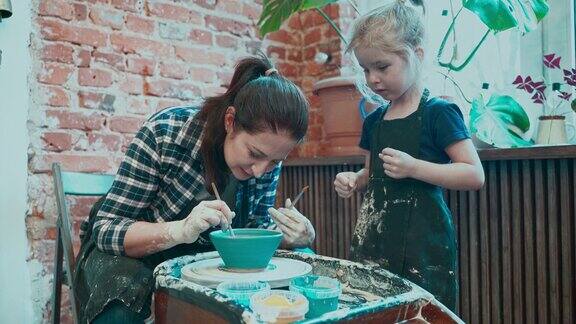 年轻的妈妈和她的小女儿在陶轮上学习陶艺在现成的碗上画画