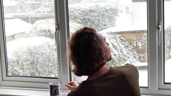 一个男人从窗框里看着雪花飘落从杯子里喝水