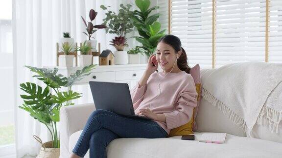 年轻迷人美丽的亚洲女性放松坐在或躺在沙发沙发或地板在舒适的位置在舒适的家庭客厅与绿色花园室内植物愉快的工作场所在家工作
