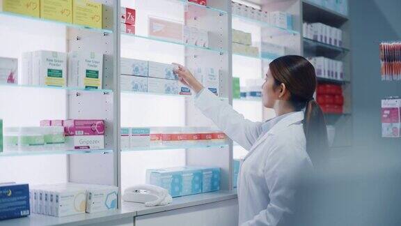 药店:美丽勤奋的亚洲药剂师的肖像穿着白大褂安排药包药盒维生素和补充药丸在柜台后面的架子上