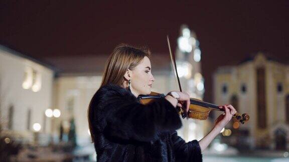 夜晚在城市建筑的背景下广场上的街道上一个浪漫的女孩穿着温暖的衣服拉着小提琴