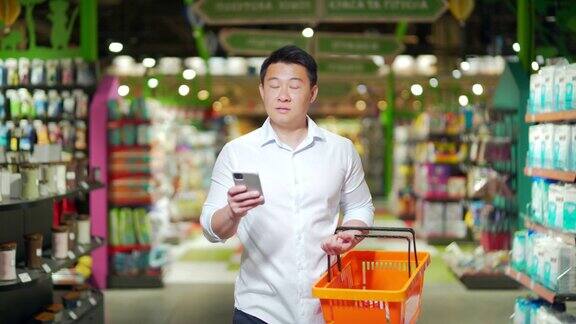 亚洲男性购物者在超市里推着购物车看着智能手机浏览智能手机搜索商品或商品清单