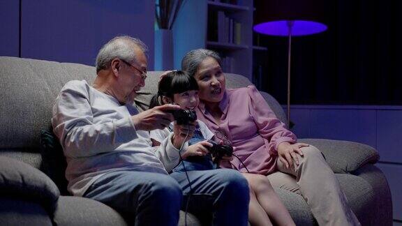 亚洲人快乐的家庭玩耍时间小女孩和爷爷奶奶一起玩电子游戏用操纵杆控制晚上在家里的客厅里开怀大笑幸福儿童幸福生活理念