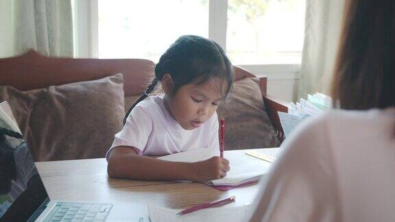 亚洲女童学生做她的家庭作业而母亲在家工作由于Covid-19大流行在隔离期间孩子在家上学父母用电脑在家工作