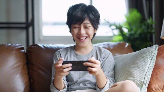 亚洲十几岁的男孩在家里客厅的沙发上玩手机游戏亚洲男孩坐在沙发上玩手机游戏