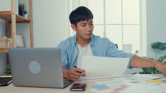 年轻的亚洲人工作与笔记本电脑坐在工作桌旁在客厅在家里