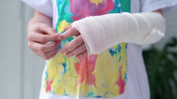 孩子在用白色石膏绷带包扎的断手手指上做按摩医疗保健女孩拿着一条断了的胳膊背景是一件五颜六色的t恤
