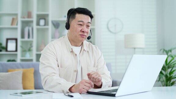 自信的亚洲商人戴着耳机在笔记本电脑上视频通话而坐在家里的办公室