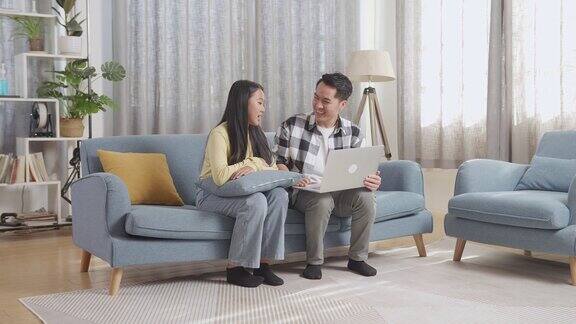 一位亚洲父亲和他的女儿坐在家里客厅的沙发上用笔记本电脑进行视频通话