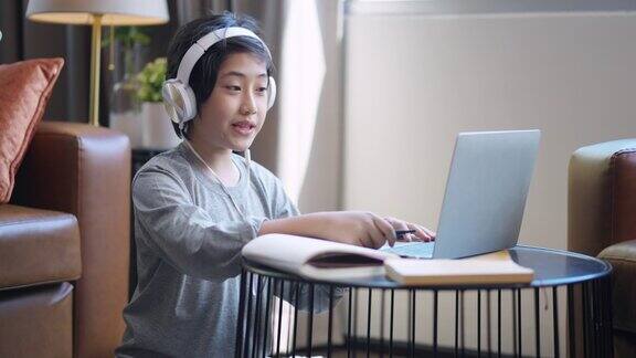 亚洲孩子男孩孩子接受电子学习学习在线课程从学校老师通过数字远程设备笔记本电脑互联网会议在客厅亚洲青少年戴着耳机使用笔记本电脑在家里学习课程