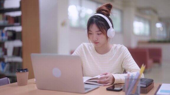 一名年轻的亚洲学生戴着耳机通过笔记本电脑进行在线学习在学校或大学图书馆安静的环境中用笔记本做笔记