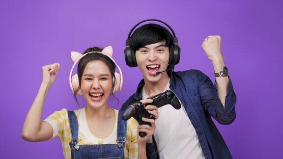 亚洲年轻夫妇玩电子游戏用操纵杆和耳机在紫罗兰色背景隔离