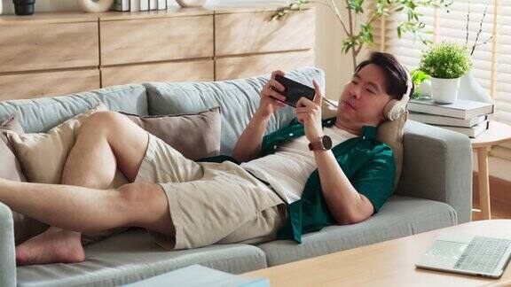 开朗的亚洲男性休闲放松玩在线多人智能手机游戏应用亚洲有吸引力的男性坐在沙发上使用智能手机玩手机游戏