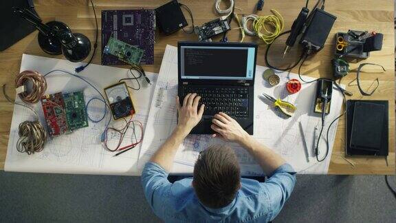 一个天才的IT技术员写代码在他的笔记本电脑坐在他的桌子上他被各种技术组件包围阳光照在他的桌子上