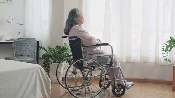 坐在轮椅上的悲伤的成熟女人看着家里的窗户