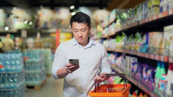 亚洲男性购物者在超市里推着购物车看着智能手机浏览智能手机搜索商品或商品清单