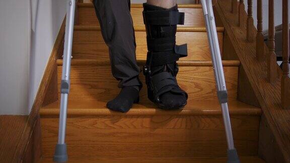 一名穿着黑色骨折靴的断腿男子试图拄着拐杖在客厅的楼梯上行走