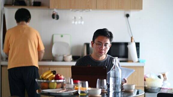 一位亚洲华人中年男子在家吃饭时使用平板电脑和文件而他的妻子在洗碗