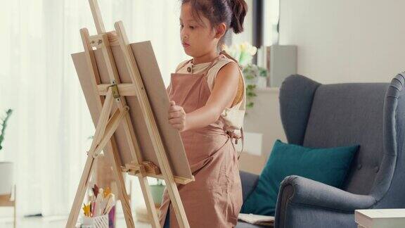 亚洲学步女孩用围裙坐沙发椅子前面的油画画布把丙烯酸色在调色板上使用调色板刀混合颜色和做抽象艺术在客厅家里