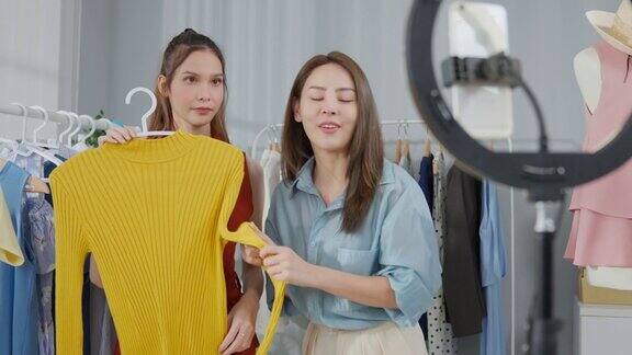 两个亚洲美女聊着镜头向家里卖衣服年轻有吸引力的商人通过虚拟在线直播工作在商店向顾客展示和销售服装产品